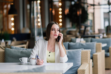 Young woman in white jacket working in cafe with papers, drinking coffee