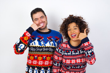 Young couple wearing Christmas sweater standing against white wall smiling doing phone gesture with hand and fingers like talking on the telephone. Communicating concepts.