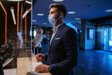 A middle-aged man in formal wear with a face mask standing on the reception of a fancy hotel and checking in.