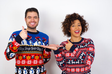 Young couple wearing Christmas sweater standing against white wall Showing palm hand and doing ok gesture with thumbs up, smiling happy and cheerful.