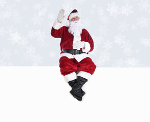 Senior man in traditional Santa Claus Suit sitting on a white wall with one hand in the air and the other on his belly. Isolated on white with copy space.