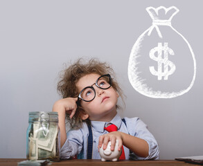 Five years old child sitting at the table with jar and piggy bank and dreams about big bag with...