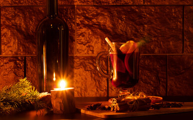 Glass Cup with mulled wine, candle, dark glass bottle, fir branch. On the Board are cinnamon sticks tied with linen thread. All in a dark interior. Background in the form of a stone wall.