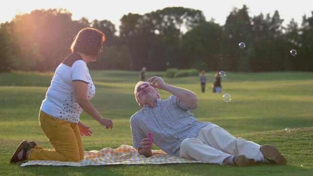 Playful senior couple blowing bubbles outdoor. Happy mature man and woman spending time together on green meadow at park.