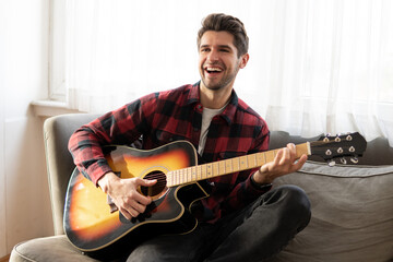 Happy young man is playing the guitar and singing sitting in the sofa 