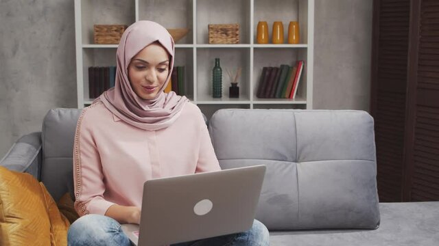 Young and attractive Muslim woman in hijab works at home using laptop computer. Middle Eastern freelancer or student on the couch.