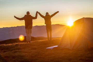 A young couple in front of the tent is holding hands and looking at the sunset at the top of the mountain. 