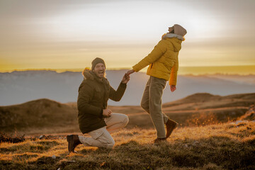 Young man making a marriage proposal to his girlfriend on the mountain peak at sunset.  - obrazy, fototapety, plakaty