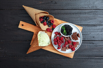 Delicious ice cream in wafer cone, chocolate and berries on black wooden table, top view