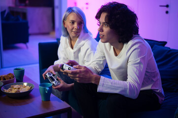 Young cheerful Caucasian husband and wife play videogames against each other at home enjoying leisure time