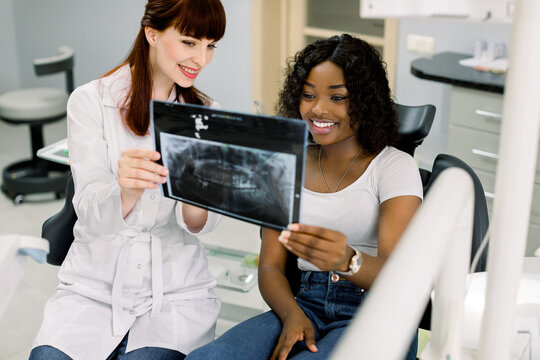 Young Caucasian woman dentist in white coat, holding x-ray image of teeth, talking with her pretty smiling young African woman patient, explaining surgical or orthodontic treatment