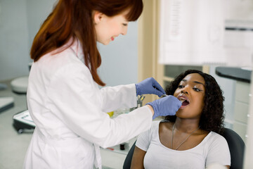 Young African female patient at dental office, opening mouth while smiling woman dentist in uniform and blue latex gloves, checking condition of teeth, using dental mirror. Oral care concept