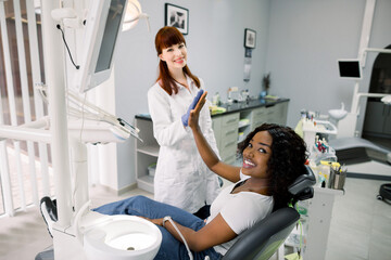 Attractive young African woman giving high five and talking to beautiful female Caucasian dentist while sitting in chair at the dentistry office. Pretty smile and successful dental treatment