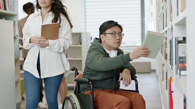 Medium long shot of handicapped Asian male person sitting in wheelchair, taking book off shelf, opening it and reading. Diverse students standing, walking in public library