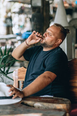 Portrait of brutal pathos man with a beard and enjoying a smoking cigar, sitting at the table. Outdoor concept.