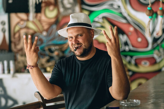 Portrait of a smiling bearded man sitting on a chair in a bar with a cigarette in his mouth and showing three fingers on both hands. Outdoor concept.
