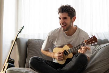 Happy young man is playing ukulele guitar and singing on the sofa at home 