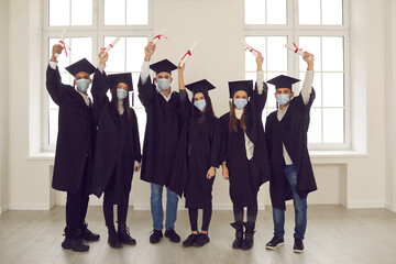 Group of successful diverse university graduates in face masks holding up their diplomas