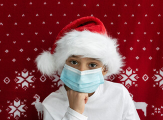 sad boy in santa hat and blue medical mask on red christmas background. copyspace for text. Holidays in new pandemic reality