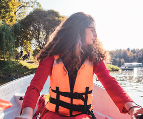 Beautiful long-haired brunette woman in an orange life jacket rowing oars while sitting in a boat....