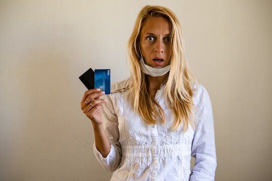 A Blond Girl Wears A Protective Medical Mask, Holds A Credit Card, On A Beige Background. The Concept Of Quarantine, Coronavirus, Shopping. Shock, Credit, No Have Money, Work After Quarantine