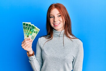 Young red head girl holding 50 israel shekels banknotes looking positive and happy standing and smiling with a confident smile showing teeth