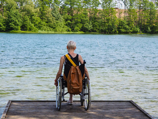A young disabled woman in a wheelchair standing on a pier on the shore of a beautiful lake
