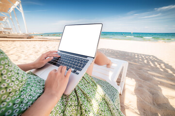Mockup image of woman using laptop with blank white desktop screen while sitting on beach chair...