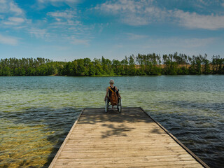 Obraz na płótnie Canvas A young disabled woman in a wheelchair standing on a pier on the shore of a beautiful lake