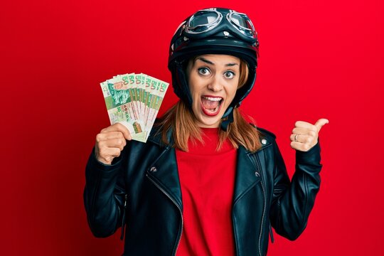 Hispanic young woman wearing motorcycle helmet holding hong kong dollars pointing thumb up to the side smiling happy with open mouth