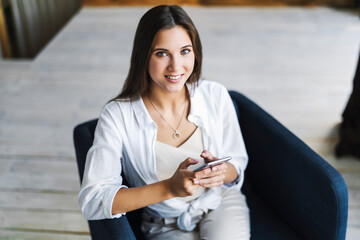 Smiling brunette in white shirt sits on an armchair, looks at camera. Portrait of business woman holding smartphone