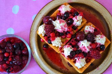 Close-up of a waffle filled with cream, cherries and berries on a pink cloth with white dots with a glass plate with more berries next to it