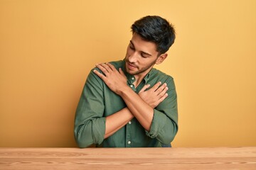 Obraz premium Young handsome man wearing casual clothes sitting on the table hugging oneself happy and positive, smiling confident. self love and self care