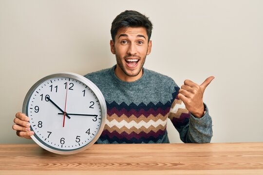 Young handsome man holding big clock pointing thumb up to the side smiling happy with open mouth