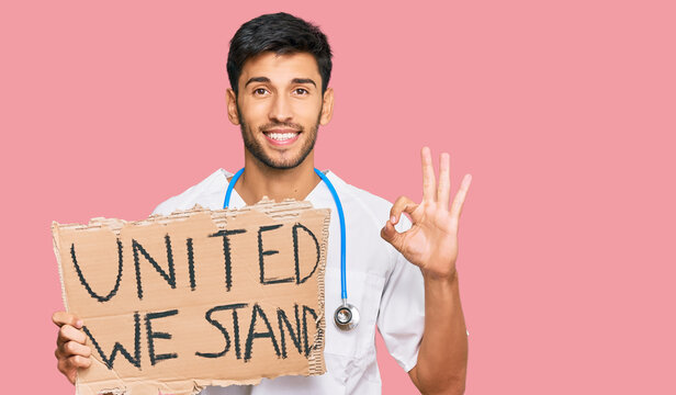 Young Handsome Man Wearing Doctor Uniform Holding United We Stand Banner Doing Ok Sign With Fingers, Smiling Friendly Gesturing Excellent Symbol