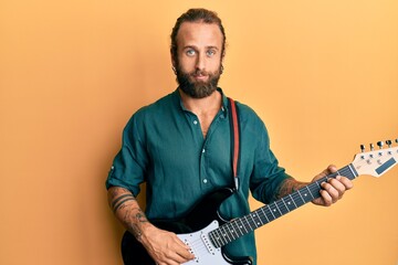 Handsome man with beard and long hair playing electric guitar relaxed with serious expression on face. simple and natural looking at the camera.