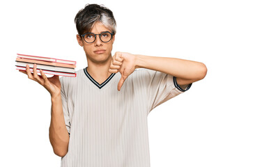 Young hispanic man holding books with angry face, negative sign showing dislike with thumbs down,...