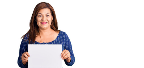 Middle age latin woman holding paper banner with blank space looking positive and happy standing...