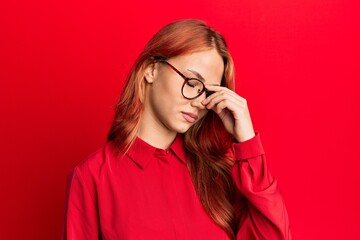Young beautiful redhead woman wearing casual clothes and glasses over red background tired rubbing nose and eyes feeling fatigue and headache. stress and frustration concept.