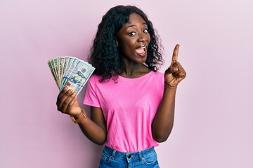 Beautiful african young woman holding dollars smiling with an idea or question pointing finger with happy face, number one