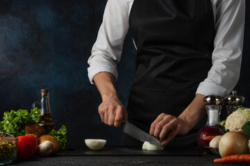The professional chef in black apron cuts with knife onion on chopped board for cooking soup on dark blue background. Backstage of preparing meal. Healthy dish. Food concept. Frozen motion. Cookbook.