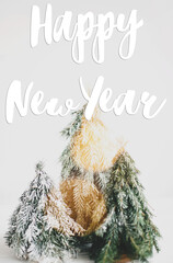 Happy New Year Greeting card. Happy New Year text handwritten on christmas little snowy trees in lights on white background, forest scene. Seasons greetings, happy holidays