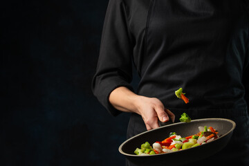 Macro shot view of chef hand throws up frying mix of colored vegetables above the pan on dark blue background. Backstage of cooking meal. Frozen motion. Food banner concept.