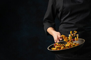 Chef in black uniform throws up frying corn with mix of colored vegetables above the pan on dark blue background. Backstage of cooking meal. Frozen motion. Food banner concept. Cookbook illustration.