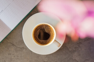 A cup of black coffee on dark stone table. Notebook and orchid in soft focus. Top view.