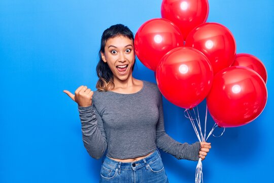 Young woman holding balloons pointing thumb up to the side smiling happy with open mouth