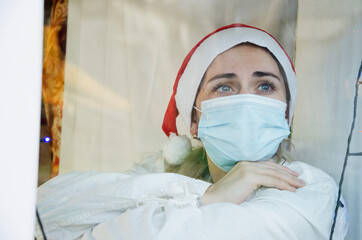 A young doctor in a Santa hat looks out the window while celebrating the New Year holidays. A doctor in uniform looks out the window. Doctor looking out the window during the coronavirus pandemic.