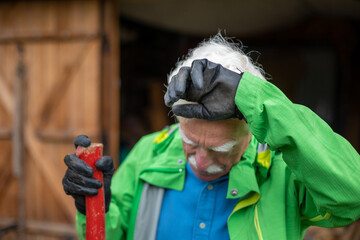 Tired senior man holding the heavy axe and wipes his forehead with fatigue.