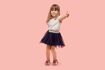 A beautiful little girl shows a thumbs up gesture and looks at the camera. In a black skirt and white t-shirt. Pink background.