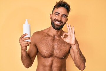 Young hispanic man standing shirtless holding sunscreen lotion doing ok sign with fingers, smiling...